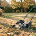Pelouse de jardin en automne avec une tondeuse arrêtée sur l’herbe sèche, des feuilles dorées éparses et une lumière fraîche de fin de journée.